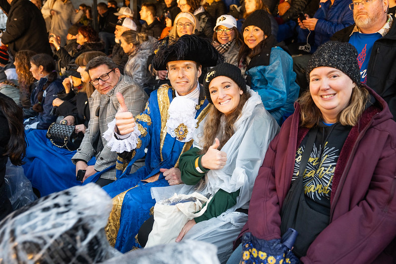 Smiling parade spectators in grandstand seating, including a performer in historic costume