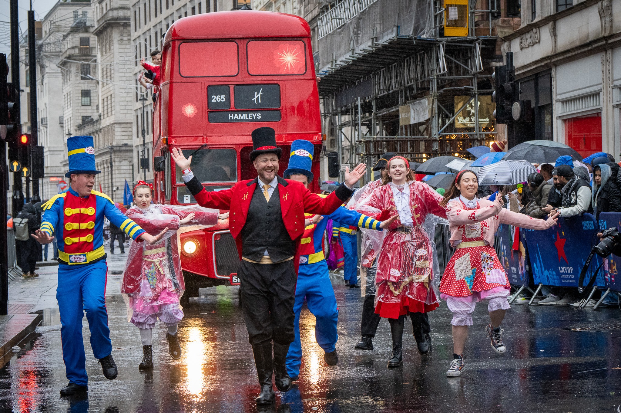 Hamleys Christmas parade with performers and red London bus in rainy city street